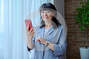 Mature business woman using smartphone, standing near window