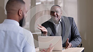 Mature businessman african boss middle age 50s leader sitting at table with laptop talking to unrecognizable man worker