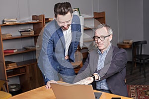 Mature businessman in blue suit is pointion on something on the laptop to his co-worker in the office