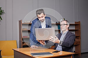 Mature businessman in blue suit is showing new project on laptop to his serious colleague in office