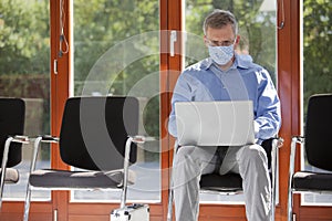 Mature businessman with face mask working with laptop in an empty waiting room of an office or hospital