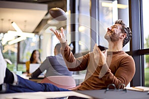 Mature Businessman With Feet On Desk In Office Talking Into Mic Of Mobile Phone Catching Football