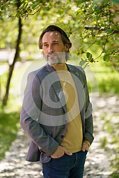 Mature businessman with grey beard wearing casual jacket and jeans spend time at park or garden looking thoughtful
