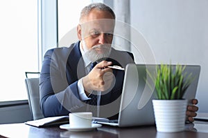 Mature businessman looking and analyzing document on laptop in his modern office at work