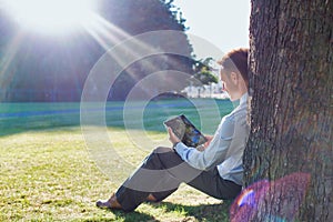 Mature businessman sitting under the tree while using digital tablet