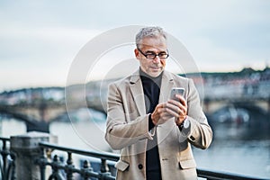 Mature businessman with smartphone standing by river in Prague city, taking selfie.