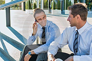 Mature businessman smiling and talking on smartphone while sitting with colleague