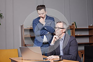 Mature businessman in suit and glasses with his younger co-worker are pensivly thinking about new project by the laptop in office