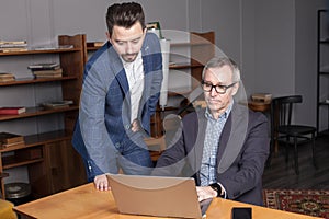 Mature businessman in suit and his pensive co-worker are diong their business work on the laptop on wooden table in the office