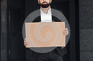 Mature businessman in suit holding blank cardboard sign standing outdoors business center, empty space