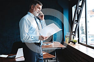 Mature businessman talking on a mobile phone standing by the window with view on city.