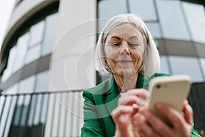Mature businesswoman holding smartphone, waiting for business partner in the city. Beautiful older woman with gray hair
