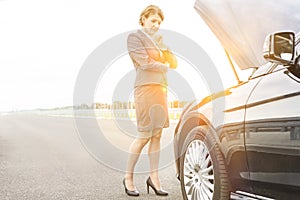 Mature businesswoman looking at breakdown car on road with yellow lens flare in background