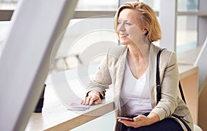 Mature Businesswoman With Passport And Boarding Pass In Airport Departure Lounge Using Mobile Phone