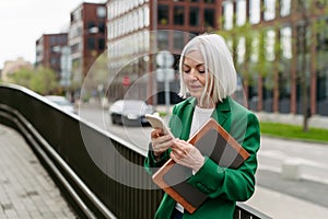 Mature businesswoman scrolling on smartphone, going home from work. Beautiful older woman with gray hair standing on