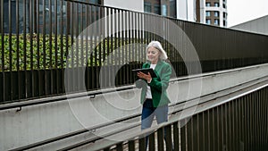 Mature businesswoman scrolling on tablet. Beautiful older woman with gray hair walking down city street.