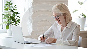 Mature businesswoman sitting at desk makes notes writing on notepad
