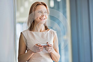 Mature businesswoman smiling while using smartphone in office hall