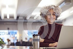 Mature Businesswoman With Travel Mug Working On Laptop In Kitchen Area Of Modern Office