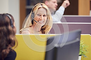 Mature Businesswoman Wearing Phone Headset Talking To Colleague In Busy Customer Services Centre