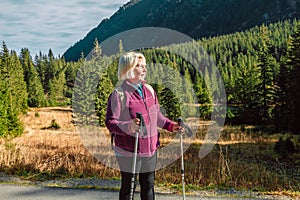 Mature caucasian blonde woman on vacation, having a hike in autumn mountains with backpack and hiking poles, enjoying