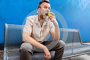 Mature caucasian man with beard eating an apple outdoors looking at camera. blue background.