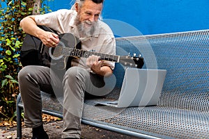 Mature caucasian man sitting on a bench learning to play the guitar with online classes using laptop.