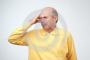 Mature caucasian man in yellow tshirtwith disgust on his face pinches nose.