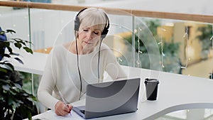 Mature caucasian woman businesswoman teacher in headphones looks at laptop screen takes notes writes report distance