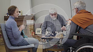 Mature Caucasian woman knitting next to men playing chess in nursing home. Lifestyle of senior people after retirement.
