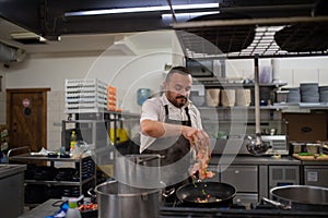 Mature chef adding chopped vehetagles to pan indoors in restaurant kitchen.