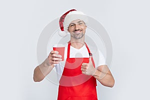 mature christmas man in red santa hat and apron hold coffee cup. thumb up