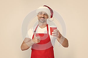 mature christmas man in red santa hat and apron hold coffee cup. thumb up photo