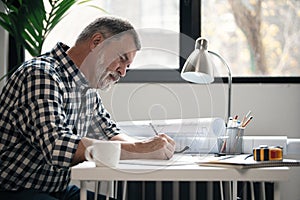 Mature confident smiling architect in casual wear working with blueprint papers and drawings in modern lightful office.