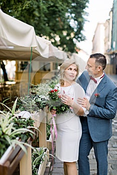 mature couple in fashionable clothes, posing near flower pots decorations at outdoors city cafe