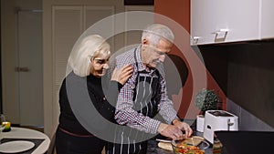 Mature couple in love making dinner. Elderly woman hugging from back husband cooking meal in kitchen