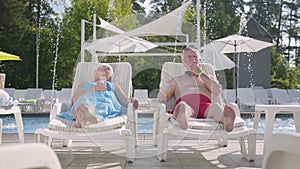 Mature couple lying on sunbeds near the pool drinking juice, talking and smiling. Happy loving family. Recreation and