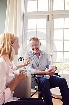 Mature Couple With Man In Wheelchair Sitting In Lounge At Home Talking Together