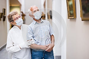 mature couple in mask protecting against covid examines paintings on display in hall of art museum