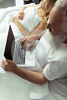 Mature couple resting in bed, man using laptop