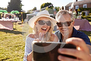 Mature Couple Sitting On Rug At Summer Garden Fete Taking Selfie On Mobile Phone