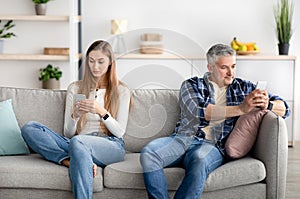 Mature couple using mobile devices, sitting apart on sofa, not paying attention to each other, playing online games