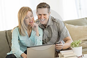 Mature Couple Using Mobile Phone And Laptop On Sofa