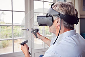 Mature Disabled Man In Wheelchair At Home Wearing Virtual Reality Headset Holding Gaming Controllers