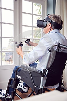 Mature Disabled Man In Wheelchair At Home Wearing Virtual Reality Headset Holding Gaming Controllers