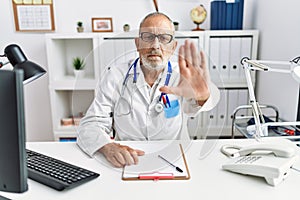 Mature doctor man at the clinic doing stop sing with palm of the hand