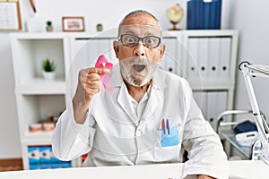 Mature doctor man holding pink cancer ribbon at the clinic scared and amazed with open mouth for surprise, disbelief face