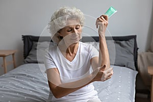 Mature elderly 60s woman applying cream on elbow in bedroom