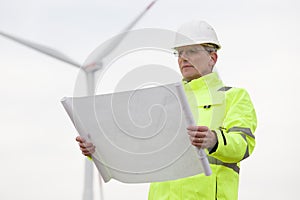 Mature engineer standing in front of a wind turbine with blueprint in his hand
