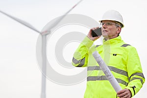 Mature engineer talking on mobile phone in front of a wind turbine with blueprint in his hand